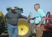 Two men facing each other in conversation and standing beside a tractor and equipment with a clear, blue sky overhead.