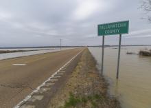 The view down a two-lane road with a wide expanse of water on each side and nearly touching the road. A road sign marks the Tallahatchie County line.