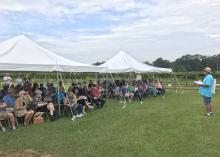 A crowd sits under tents as a speaker addresses them.