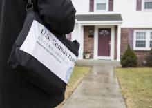 A person carrying a black bag marked with census information approaches the front door of a house.