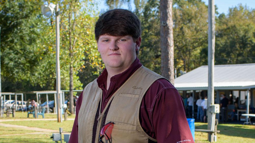 A teenage boy stands outside with a firearm resting on his shoulder.