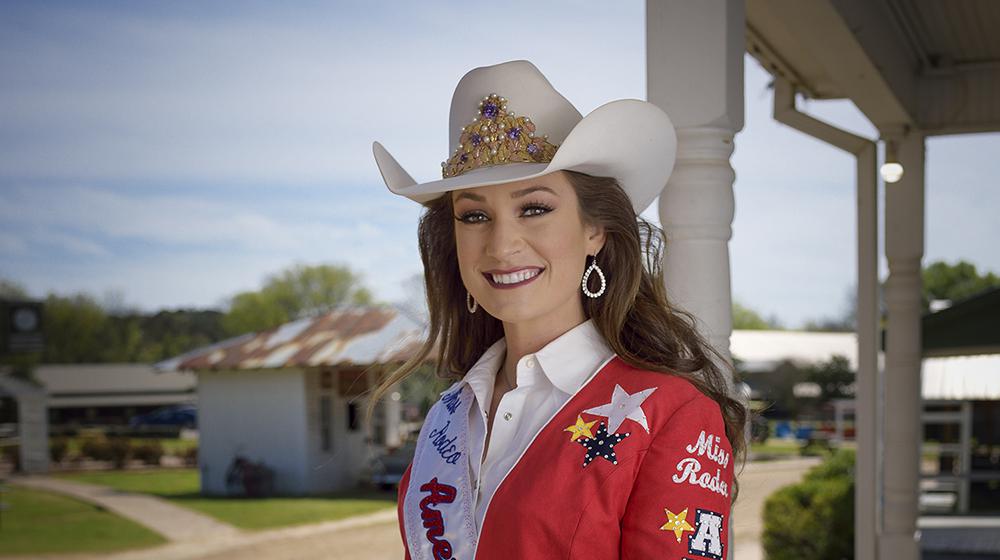 Taylor McNair, former Hinds County 4-H'er A young woman wearing a white cowboy hat and a red embroidered blazer stands on a wooden deck.