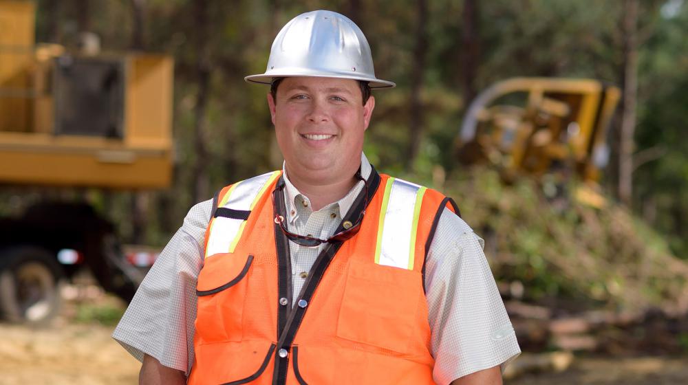 Drew Massey, logger A young man with a metal construction hat and bright orange vest stands in front of his work site with his hands tucked into his blue jeans.