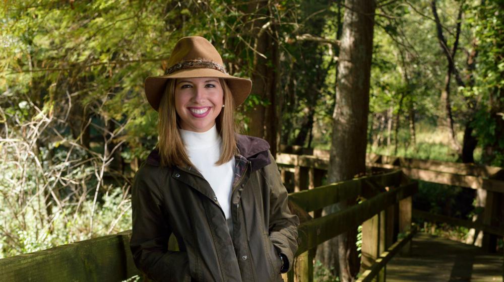 Ashley Ward, Former 4-H member A young woman with brown hair wearing a tan hat with a green jacket and khaki pants leans against a wooden railing.