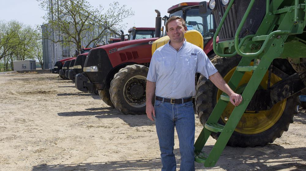 A man wearing a blue collared shirt rests his hand on a green tractor with several red tractors parked behind him.