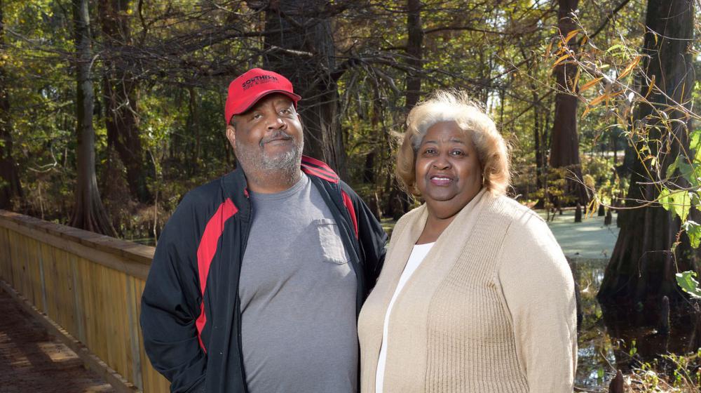 Mattie and Willie Williams, 4-H volunteers A couple, a man with a black and red jacket and a woman with a tan cardigan, stands next to a wooden railing located outside with trees and water in the background.