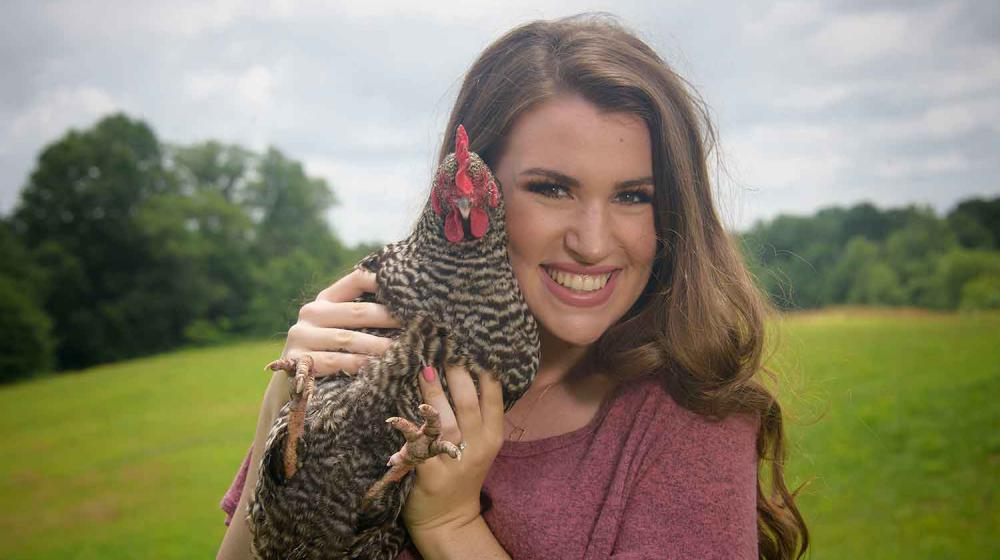 Chicken Care A young woman with brown hair and a pink shirt stands smiling while holding a black and white speckled chicken next to her face.