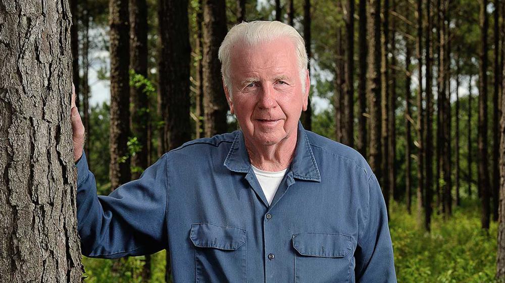 A white-haired male wearing a denim shirt and jeans stands beside a tree and smiles.