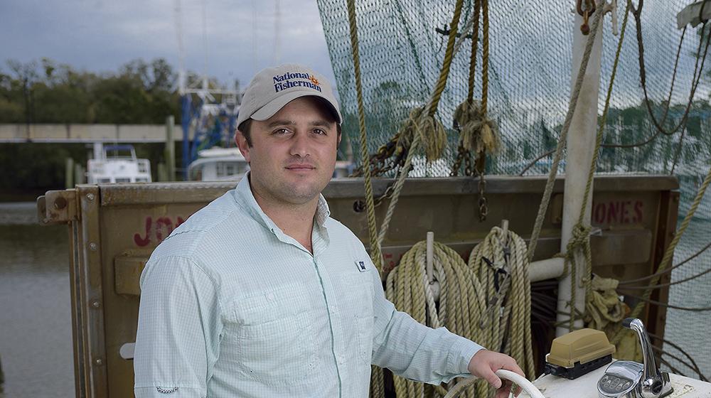 A man wearing a blue collared shirt stands in a boat with his hand on an old white steering wheel.