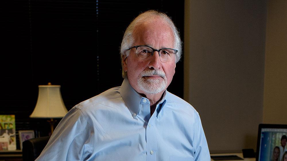 A mean wearing a blue dress shirt leans against a desk.