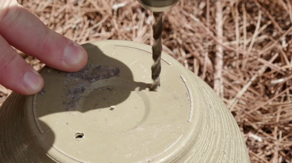 Fingers steady an upside-down flower pot as a drill bit pierces the bottom to make drainage holes. 