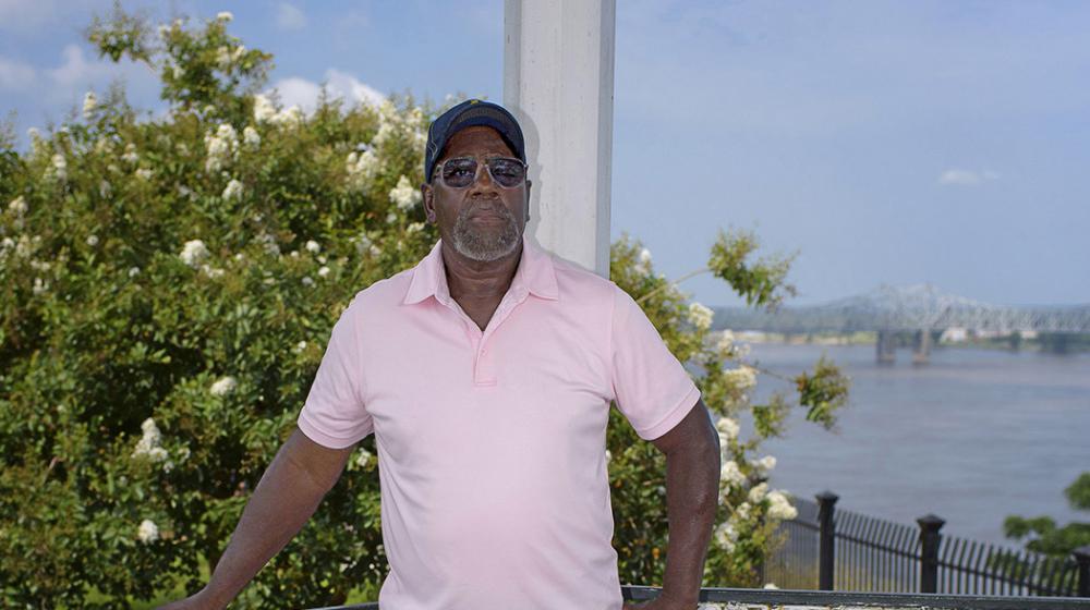 A man stands on a porch with a white crepe myrtle behind him and the Mississippi River and bridge in the distance.