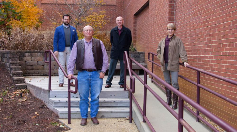 Three men and one woman standing, spaced out in front of a red brick building.