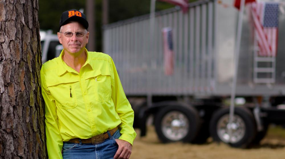 A man wearing a bright yellow button-up shirt leans against a large truck and rests one hand in his blue jean pocket.