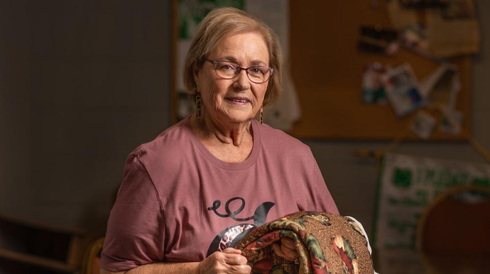 An older woman smiles while holding a quilt.