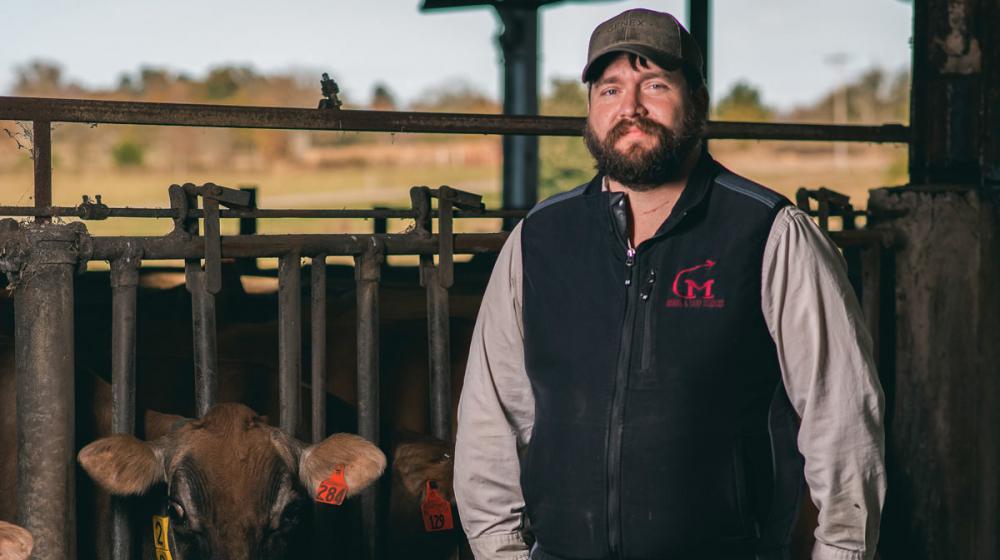A man gazes off to the left as the sun sets behind him and two cows look at him from the opposite side of the fence he is leaning on.