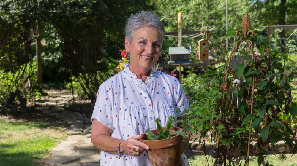 A woman wearing a white shirt with red and blue stars holds a potted plant in a garden.