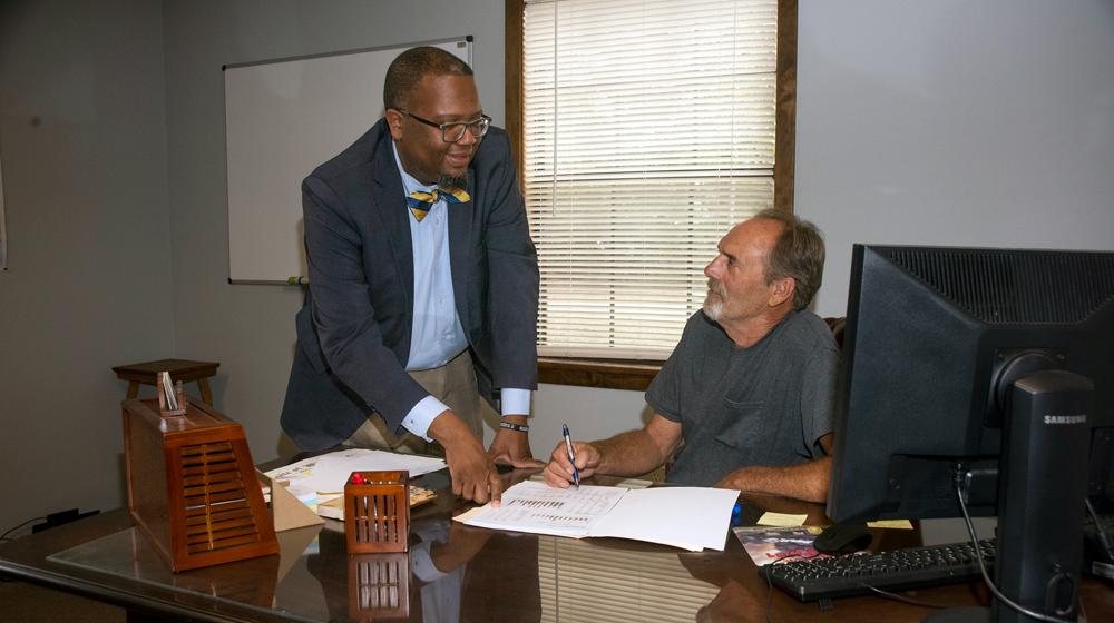 A standing Black man points to a paper on a desk that a seated white man with a pen also points to.