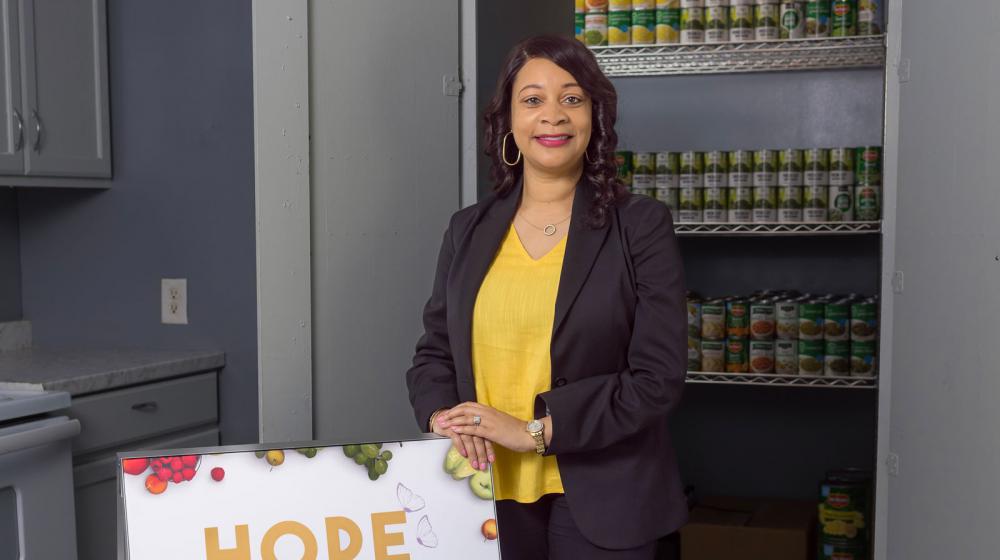 A professionally dressed woman standing in front of many rows of food cans and beside a sign that lists “HOPE.”