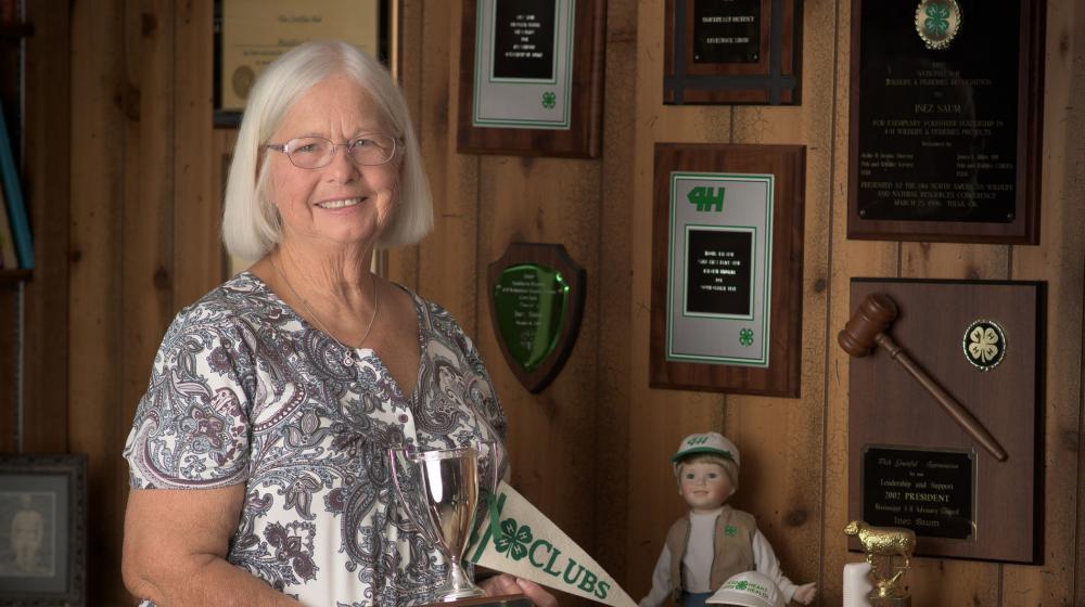 A woman, smiling, holding a 4-H flag and 4-H trophy and standing in front of several 4-H plaques affixed to the wall.