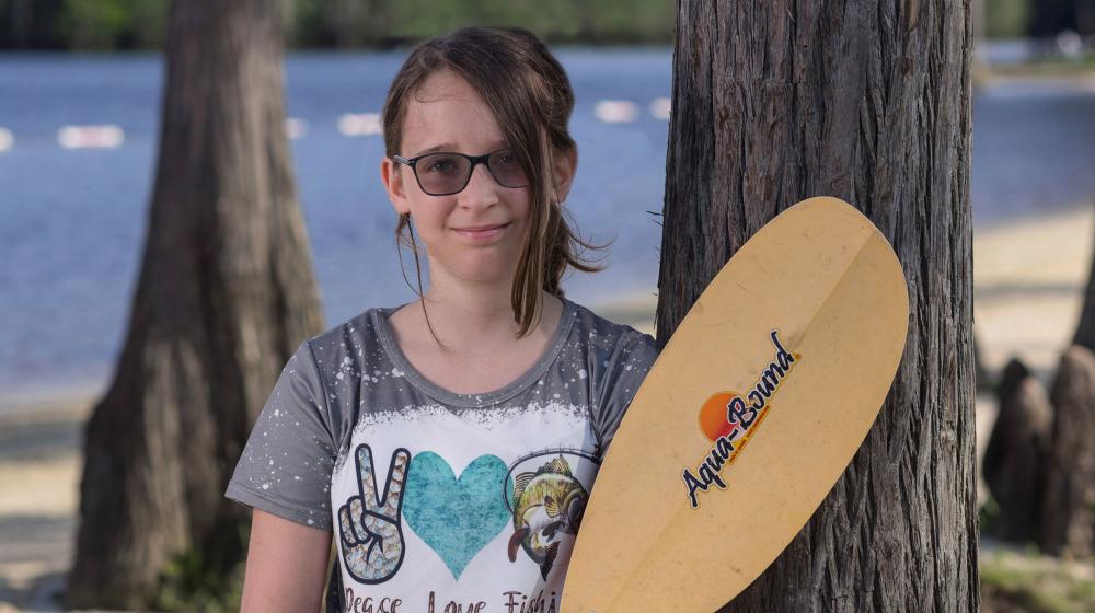 A teen girl smiling, wearing sunglasses and holding a rowing oar.