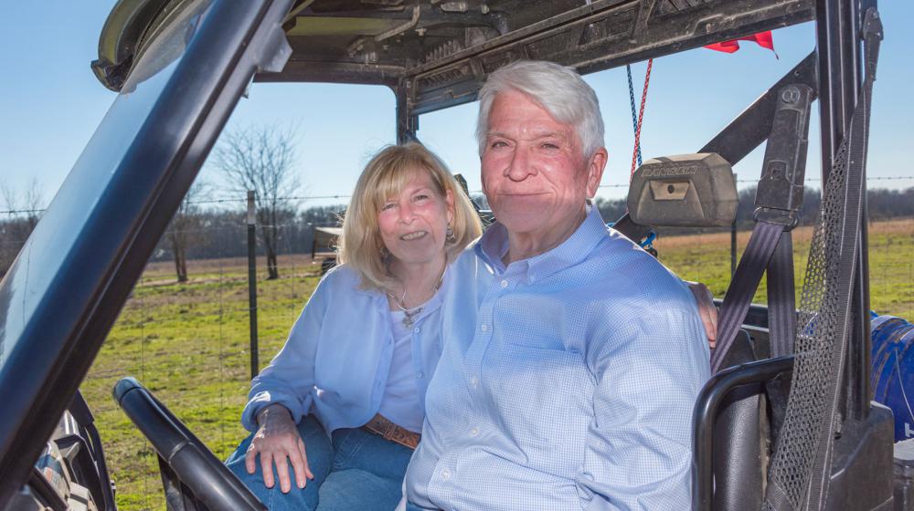 A woman and man seated in a side-by-side with a cattle field stretching behind them.