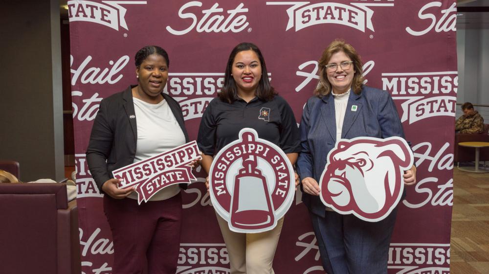 Three women standing in front of a maroon and white Mississippi State University banner.
