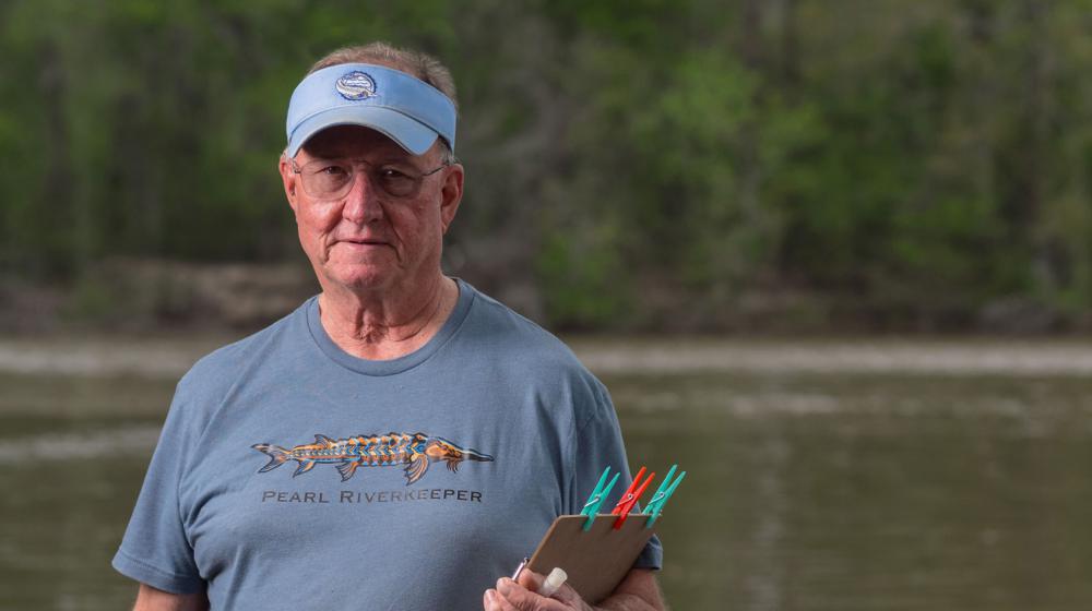 A man with a blue visor and T-shirt listing “Pearl Riverkeeper” and holding a clipboard in front of a waterway.