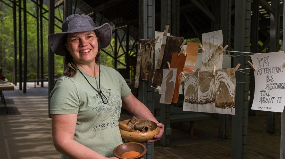 A smiling woman, holding a bowl of rocks in one hand and a bowl of sandy colored dirt in the other, standing in from of paintings hung on a line to dry.