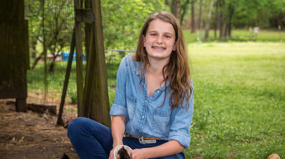 A girl wearing denim smiles as she feeds her chickens.