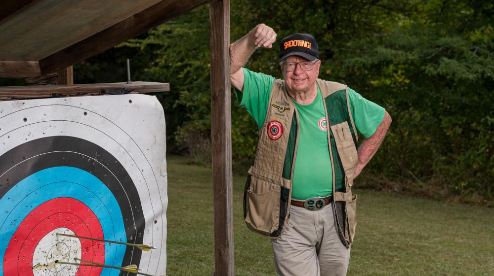 A smiling man wearing a vest and green shirt, standing beside an archery target with three arrows near the bullseye.