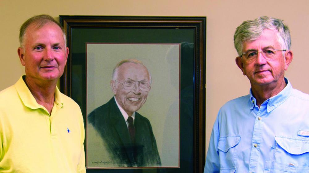 Two men, one in a yellow polo and the other in blue, stand in front of a portrait of a man