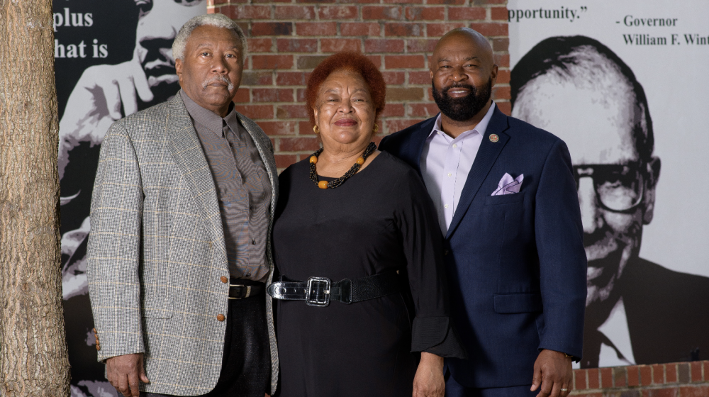 An older man and woman with a younger man stand smiling in front of a sign honoring Sadye Weir.