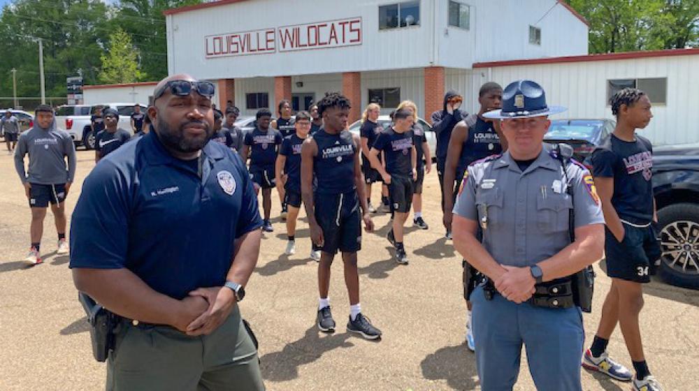 Two officers stand in front of a group of young men spaced apart for workouts in a parking lot.