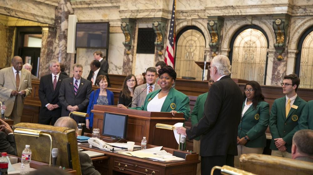 A young woman wearing a dark green blazer stands behind a podium while several people stand looking at her. 