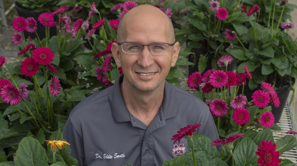 A smiling man with a polo listing his name as “Dr. Eddie Smith” is surrounded by colorful flowers.