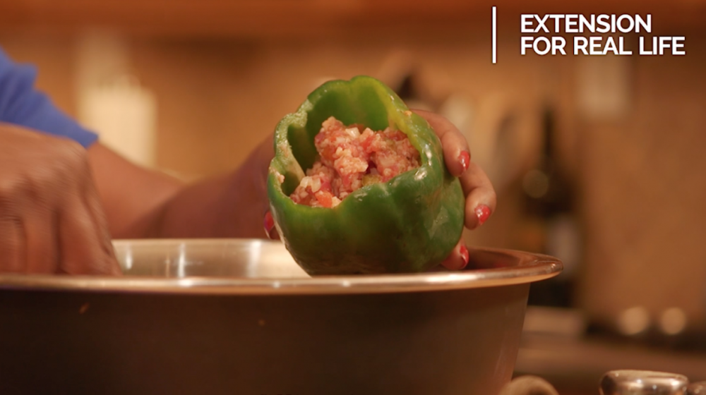 Hands spoon a ground meat and riced cauliflower mixture into a raw green bell pepper held over a stainless steel bowl. 