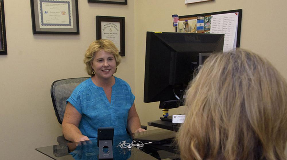 A woman wearing a blue shirt sits behind a computer and smiles at a woman seated across from her.