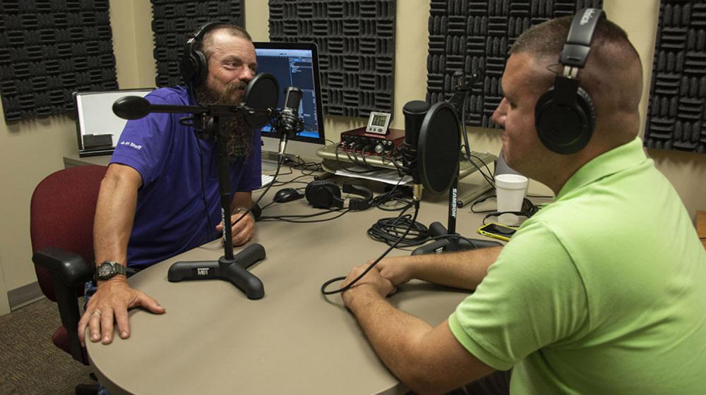 A man in a purple shirt speaks into a microphone as a man in a green shirt, also in front of a microphone, watches.