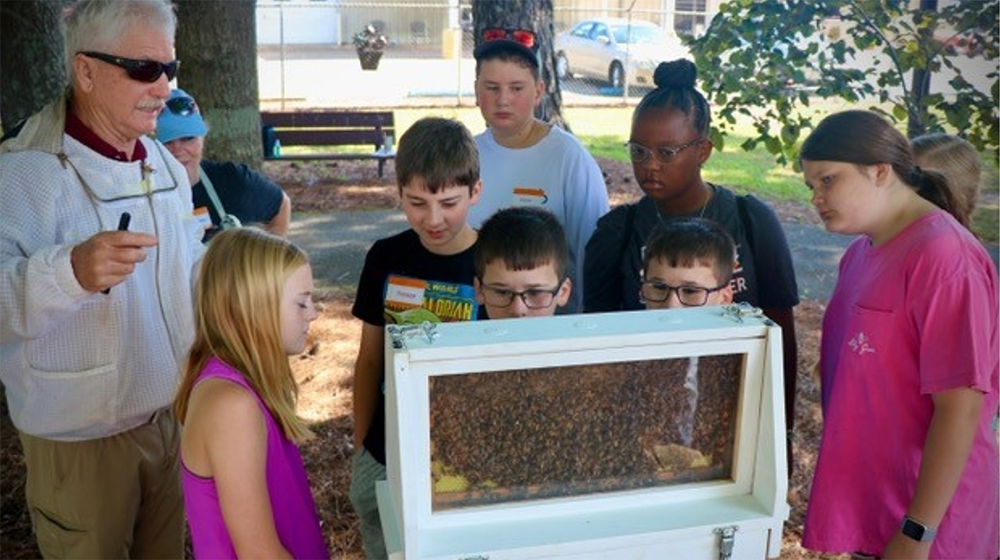 A man with a group of young people standing in front of a white manufactured beehive.