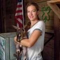 A blonde woman in a white shirt and ponytail stands in front of a wooden wall and holds a brown, black, and white baby goat. 