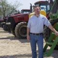 A man wearing a blue collared shirt rests his hand on a green tractor with several red tractors parked behind him.