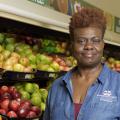 A woman wearing a blue collared jacket stands in the fruit aisle of a grocery store.