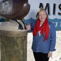 A smiling woman with blonde hair, a red scarf, a denim shirt, and black pants rests her arm on the side of a “Welcome to the Mississippi Gulf” sign.