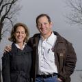 A man stands next to a woman with his arm around her shoulders while they both smile at the camera; a lake rests in the background.