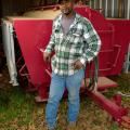 A man wearing a green and white plaid shirt, blue jeans, and a brown baseball cap stands in front of a bright red machine parked inside a metal building.  