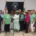 A group of 14 men and women stand on either side of a woman wearing a bright green hat and holding up an award.