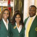 Two young women and one young man wearing green blazers stand on a marble floor.