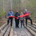 Four people stand behind a recently cut large red ribbon on a new bridge over a body of water.