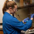 A young woman wearing safety goggles and a blue lab coat working in a lab.